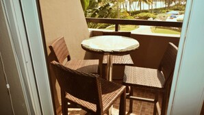 A small balcony with wicker chairs and a glass table, overlooking a tropical landscape with palm trees and a view of the ocean.
