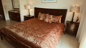 A large bed with a patterned bedspread, two bedside tables with lamps, a painting on the wall, and a bathroom in the background.