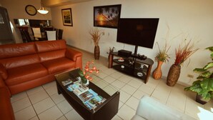 A living room with a brown leather sofa, a coffee table with magazines, a flat-screen TV on a stand, and several potted plants.