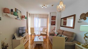 Ein Wohnzimmer mit Fernseher, einer Couch, Sesseln, einem Couchtisch und einer Deckenleuchte.