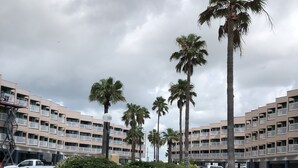 A parking lot with several cars, surrounded by palm trees and a multi-story building with balconies. - Steps from the Beach, sun, surf and boardwalk! (Corpus Christi)