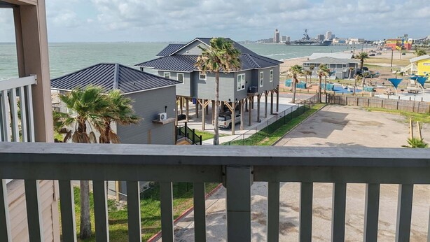 A beachfront property with elevated structures, a sandy beach, and a distant city skyline. - Steps from the Beach, sun, surf and boardwalk! (Corpus Christi)