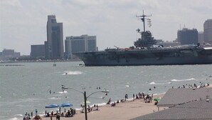 A large naval ship docked near a beach with people and buildings in the background. - Steps from the Beach, sun, surf and boardwalk! (Corpus Christi)