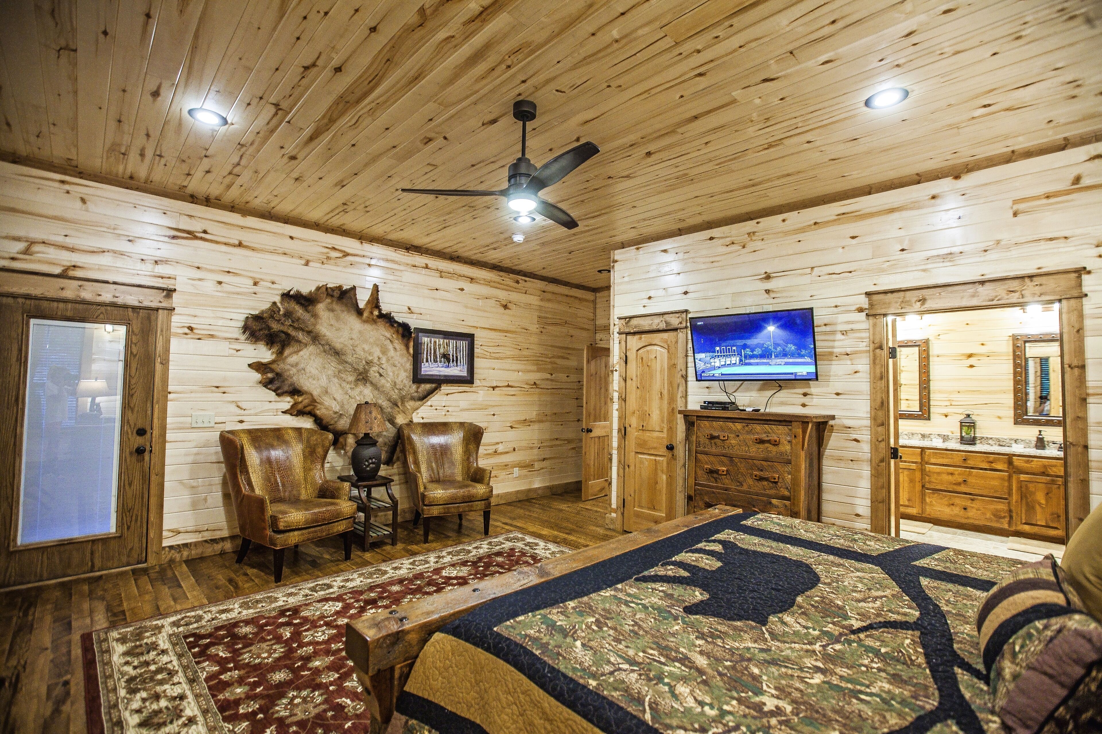 A log cabin room with a bed, two leather chairs, a television, and a mounted animal hide.