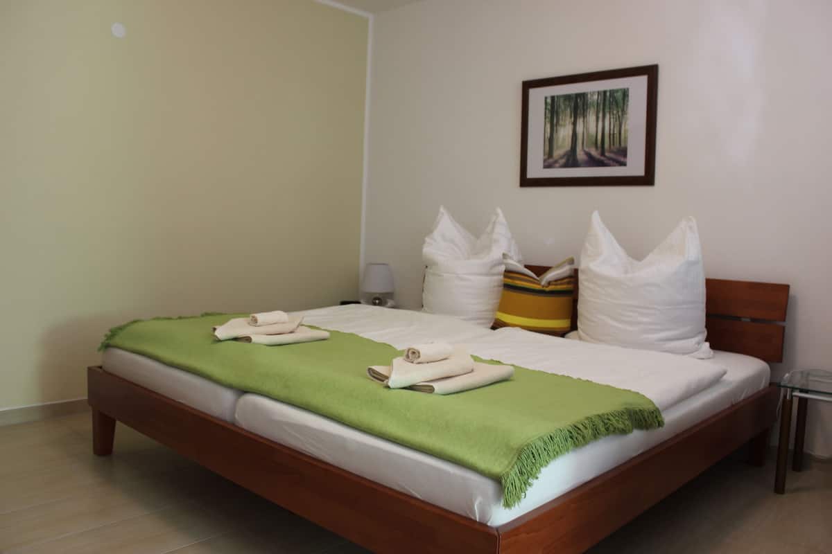 A neatly made bed with a green throw blanket and white pillows, a bedside table with a lamp, and a framed picture on the wall.