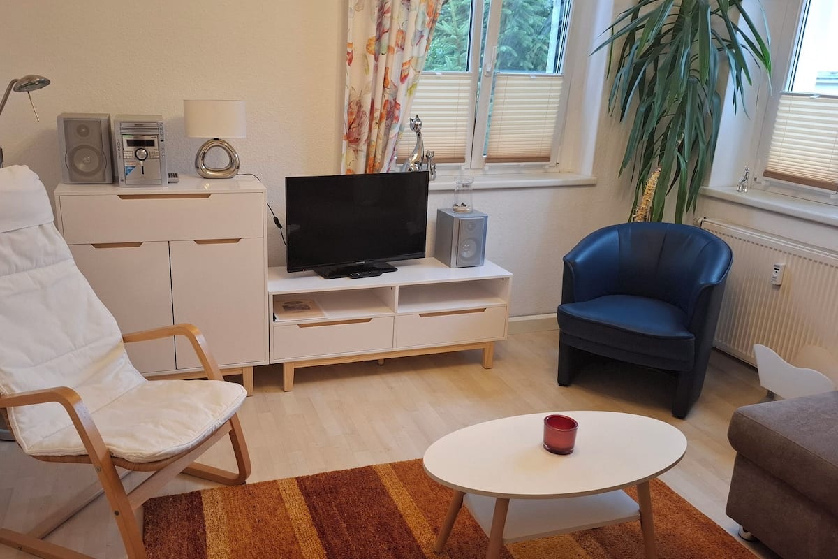 A modern living room with a white entertainment unit, a flat-screen TV, a blue armchair, a white armchair, and a small round table with a red cup.