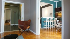 Een moderne woonkamer met een bruine leren fauteuil, een eethoek met witte stoelen, en een keuken met blauwgroene muren en witte kastjes.