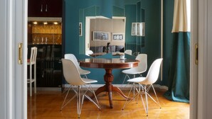 Een eethoek met een ronde houten tafel omringd door vier witte stoelen, een keuken met glazen en een koelkast, en een muur in teal met een raam en gordijnen.