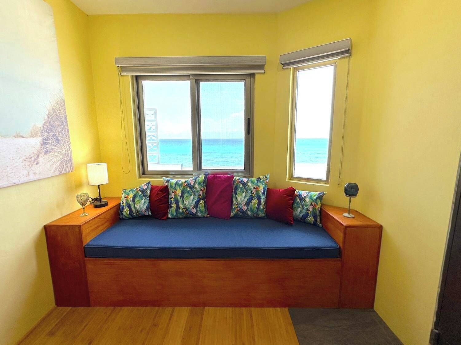 A room with a bed against a wall, two windows with a view of the sea, a lamp on a bedside table, and a painting on the wall.