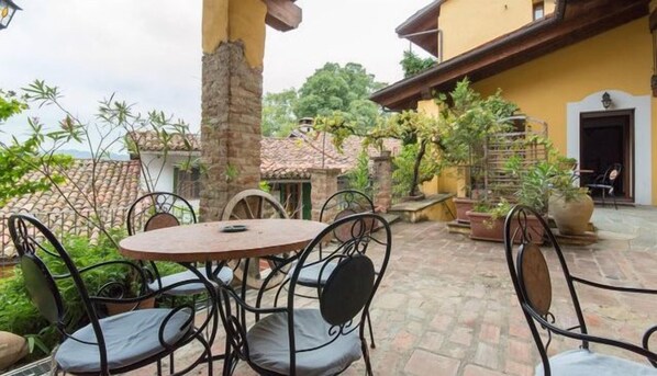 Un patio con una mesa redonda y sillas de metal, rodeado de plantas en macetas y un muro de piedra.