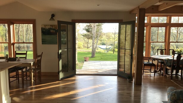A spacious room with wooden floors, a dining area with a table and chairs, and large windows offering views of a garden.