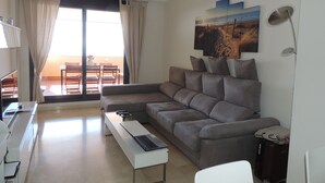 A modern living room with a grey sofa, a white coffee table, and a laptop. There is a dining area with chairs and a table visible through a large sliding door. -  Marina de Casarès Dona Julia Golf Andalusia 29690 ES (Casarès costa-malaga andalousie)