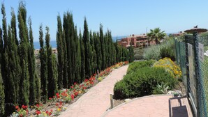 A paved pathway lined with tall cypress trees and colorful flowers, leading to a building with a view of the sea. -  Marina de Casarès Dona Julia Golf Andalusia 29690 ES (Casarès costa-malaga andalousie)