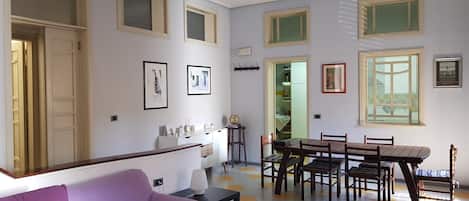 Una sala de estar con un sofá morado, una mesa de comedor con sillas y una cocina visible a través de una puerta abierta.