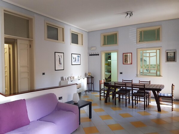 A living room with a purple sofa, a dining table with chairs, and a kitchen area visible through an open door.