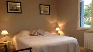A neatly made bed with striped pillows, two framed pictures on the wall, a small table with a lamp, and a window with a view of greenery. - Le Pressoir: Spacious vacation rental, surrounded by nature, forest 50 meters away, Pornic 20 minutes away (Chaumes-En-Retz)
