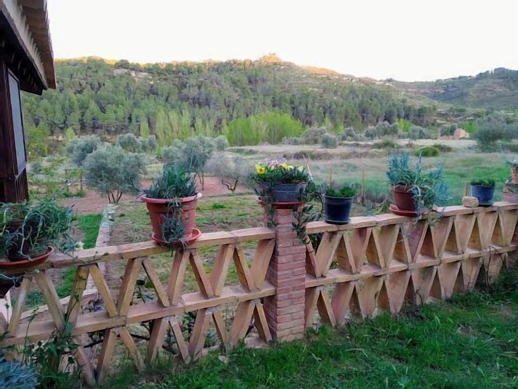 A wooden fence with potted plants, overlooking a hilly landscape with trees and shrubs.