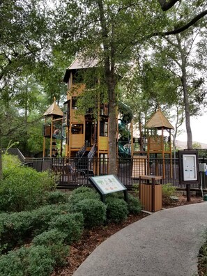 Explore a playground with a multi-level play structure and climbing elements, surrounded by lush greenery and a paved walkway.