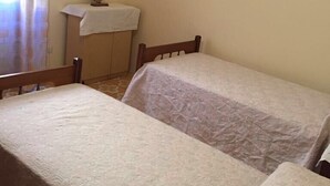 A room with two single beds, a small table with a white cloth, and a wall-mounted shelf with a framed picture.