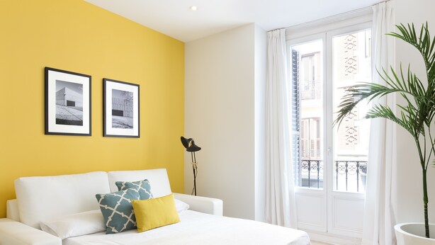 A hotel room with a yellow accent wall, a bed with white bedding, a potted plant, and two framed pictures on the wall. - CARRETAS APARTMENTS MADRID 102 (MADRID)