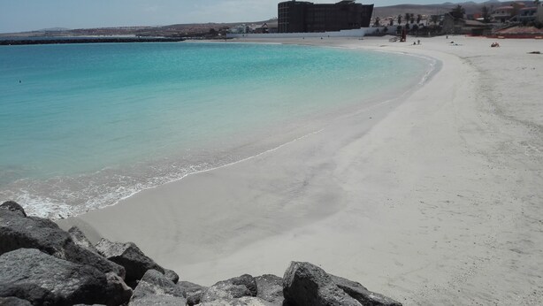 Une plage à l’eau turquoise et limpide, bordée d’un rivage sablonneux et ponctuée de formations rocheuses.