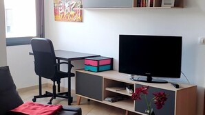 Un salon moderne doté d’un téléviseur à écran plat, d’un fauteuil de bureau noir, d’un bureau en bois avec des étagères et d’une fenêtre avec des stores.