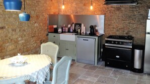 Una cocina rústica con paredes de ladrillo visto, techo de madera y estufa a gas. Hay una mesa de comedor con mantel blanco y sillas de mimbre.