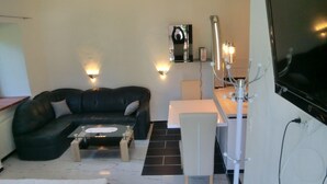 Un salon moderne avec un canapé en cuir noir, une table basse en verre, une table à manger blanche avec une chaise, et un téléviseur fixé au mur.