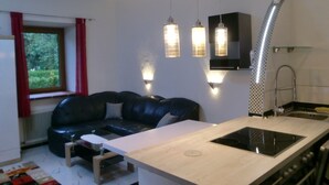 Un espace de vie compact comprenant un coin cuisine, un canapé en cuir noir, une petite table en bois et une fenêtre ornée de rideaux rouges.