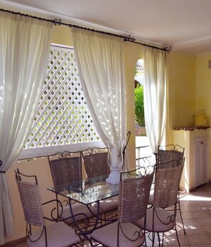 Mesa com tampo de vidro, cadeiras de vime, cortinas brancas e veneziana de treliça.