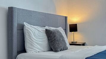 Una cama bien hecha con almohadas blancas y grises, un cabecero gris y una mesita de noche con una lámpara.