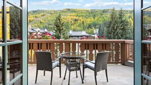 A balcony with wicker chairs and a glass table, overlooking a forested valley with autumn foliage and houses.