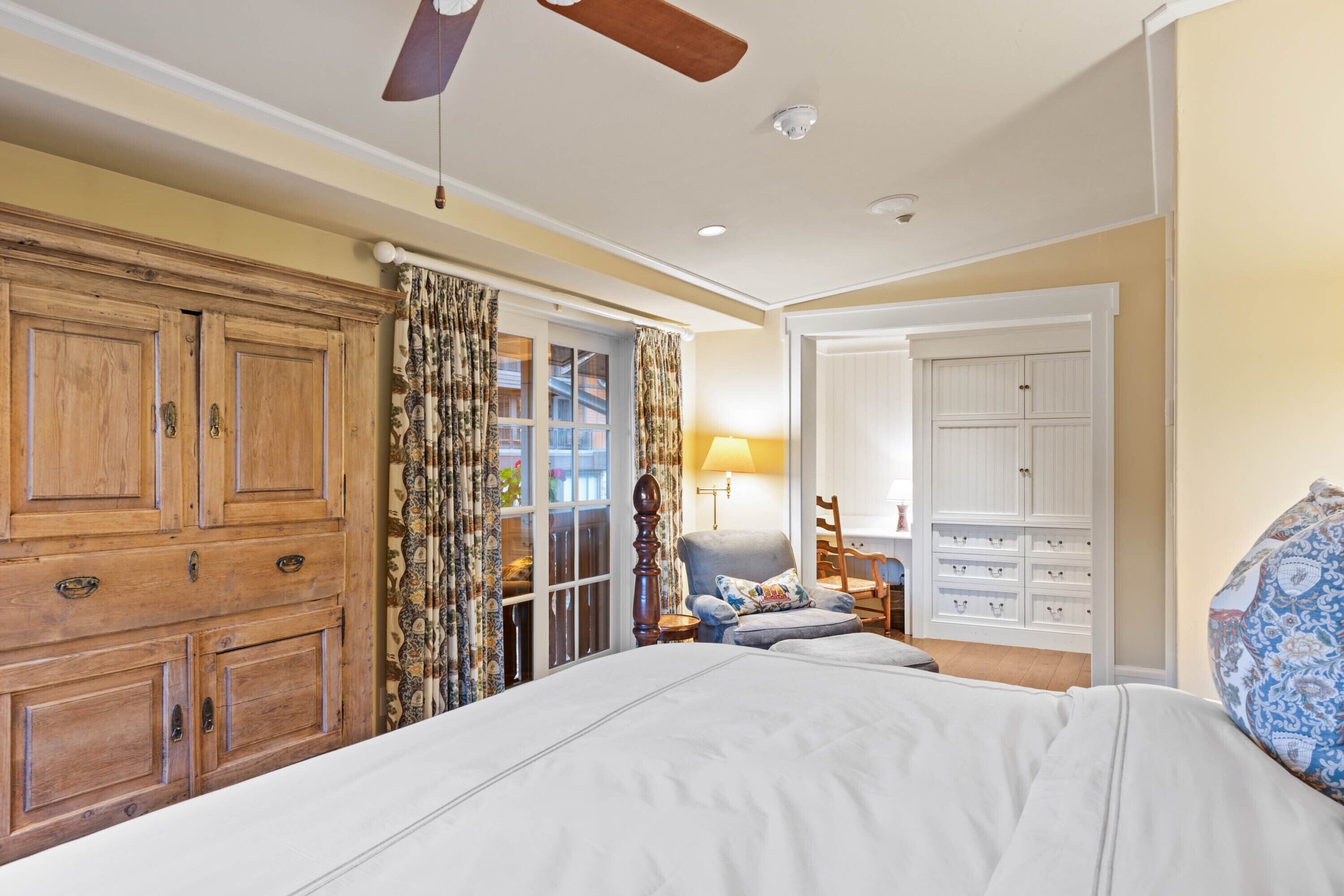 A bedroom with a large bed, wooden wardrobe, a chair, and a desk with a lamp.
