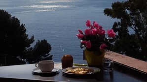 Une vue sur la mer depuis un balcon, avec une table dressée pour le petit-déjeuner, comprenant une tasse, du pain et un vase rempli de fleurs.