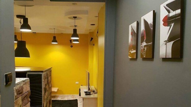 Un couloir étroit doté d’un plancher en bois, d’un mur en pierre d’un côté et d’un mur jaune de l’autre. Trois suspensions sont accrochées au plafond, et trois œuvres d’art encadrées ornent le mur jaune.