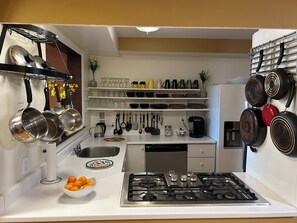 A kitchen with a gas stove, hanging pots, a coffee maker, and a bowl of oranges. - Spacious 2 Bedroom Mid-Downtown Apartment. Fully Furnished & Equipped. (St. John's)