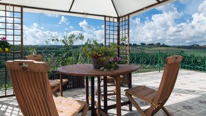 Een overdekt terras met een tafel en stoelen, een bloemenmand, en uitzicht op het landschap.