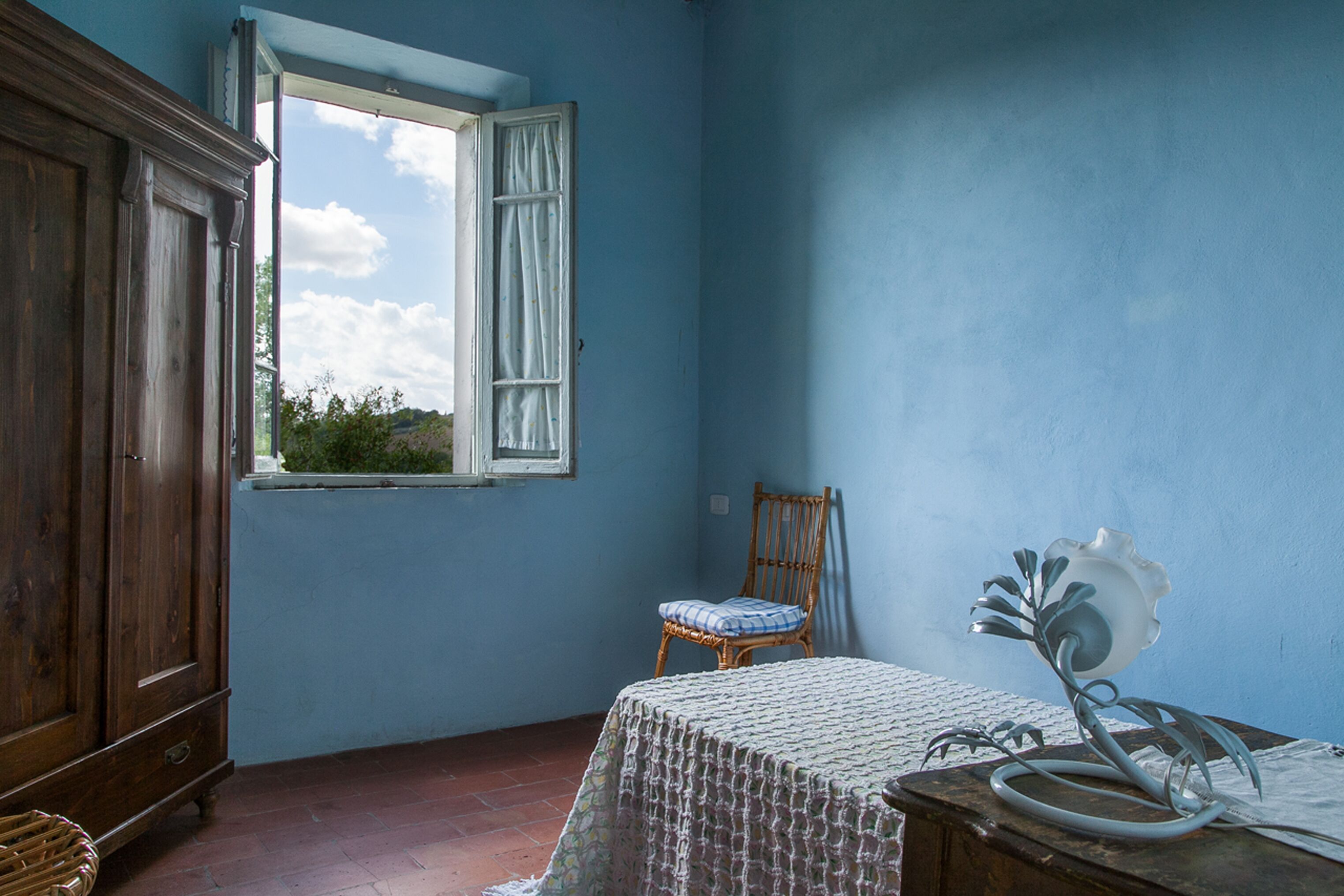 Una habitación con un armario de madera, una cama con colcha estampada, una silla de mimbre, un objeto decorativo de metal sobre una mesa y una ventana con vistas a la vegetación.
