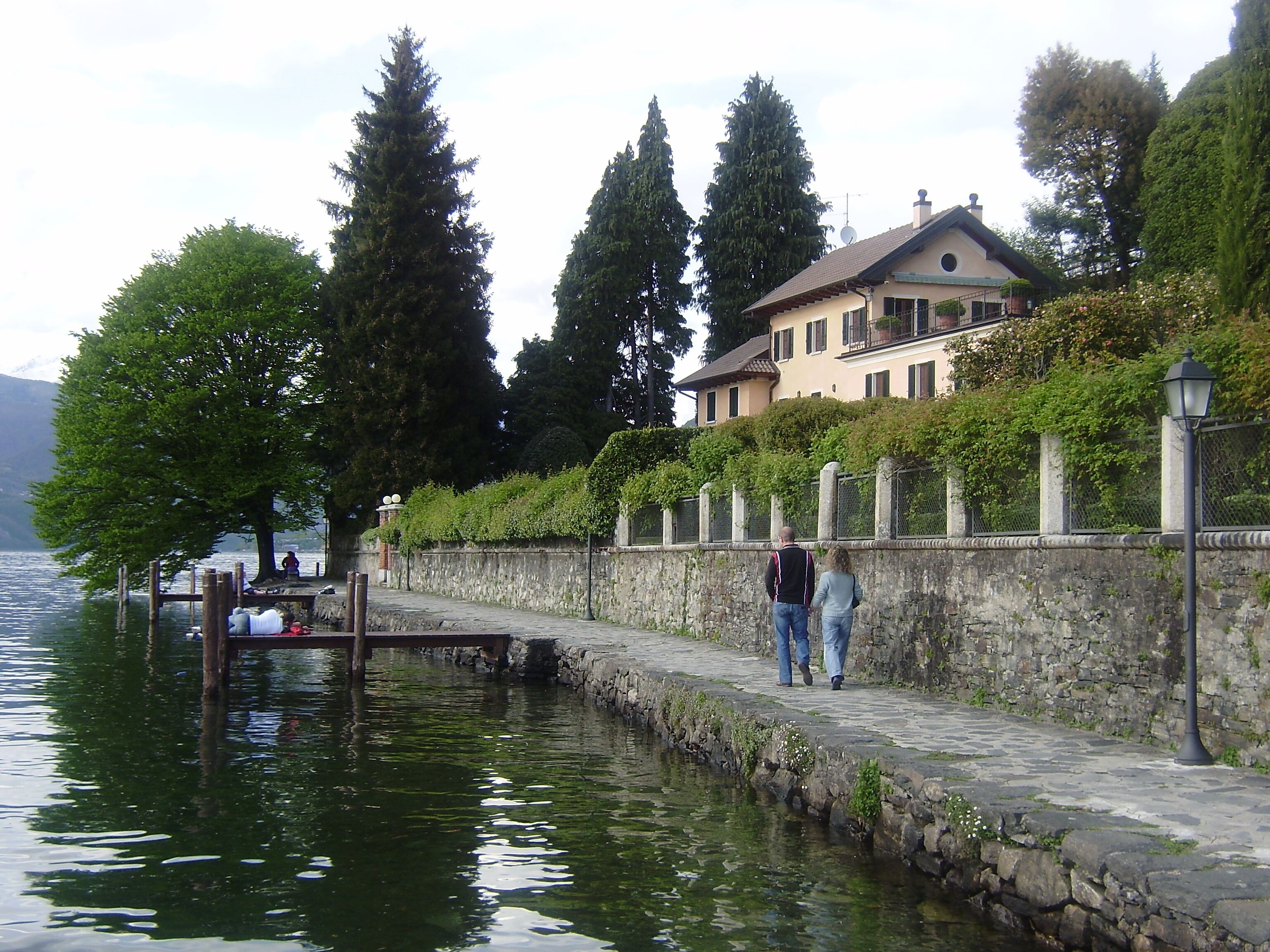 A lakeside walkway with a stone wall, a wooden dock, and a house with a balcony.