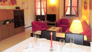Un salon avec un téléviseur, un fauteuil rouge, une table à manger dressée pour deux personnes et un buffet aux portes vitrées.