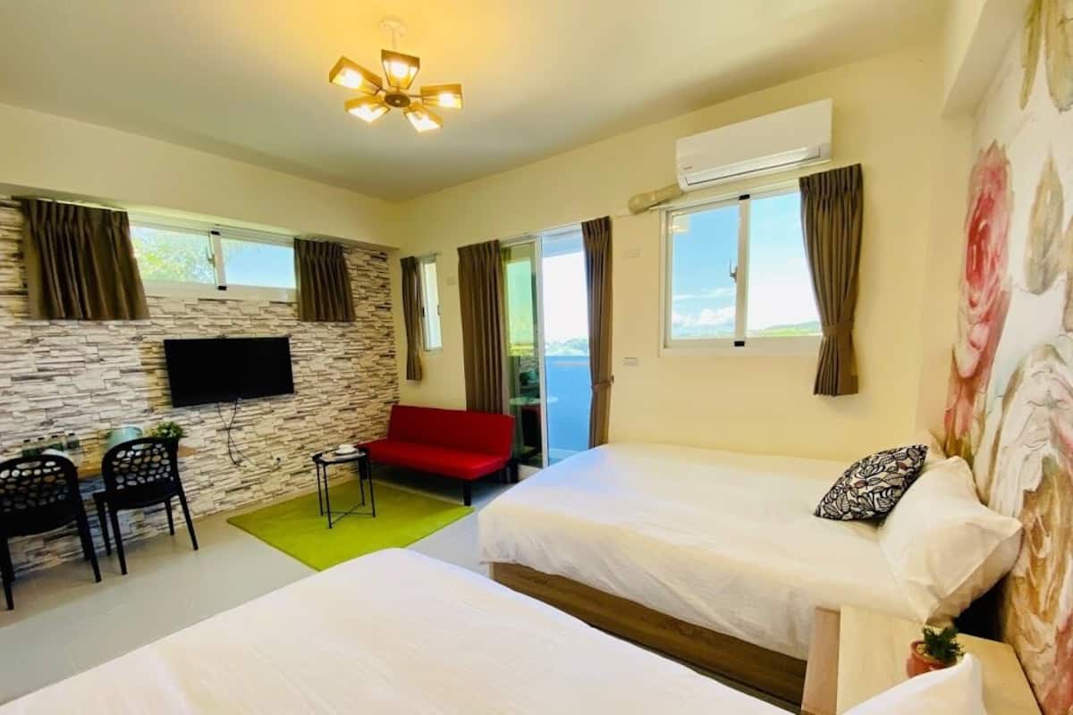 A hotel room with two beds, a TV mounted on a stone wall, a red sofa, and a view of the outdoors.