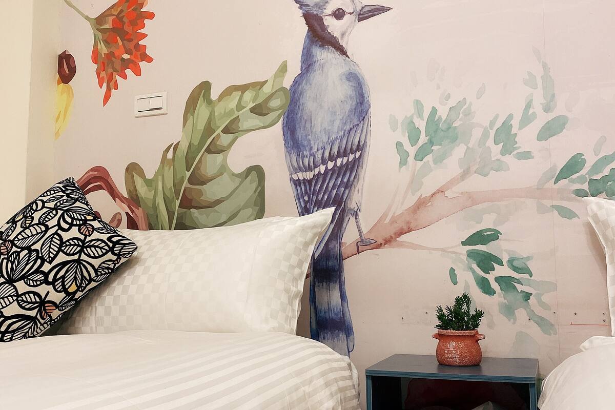 A bedroom with a bed, a nightstand, a potted plant, and a wall mural of a bird and foliage.