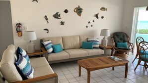 Un soggiorno con un divano color beige, due poltrone, un tavolino in legno e una sedia in vimini. Sono presenti decorazioni a parete tra cui stelle marine e un cavalluccio marino. Un estintore è fissato al muro.