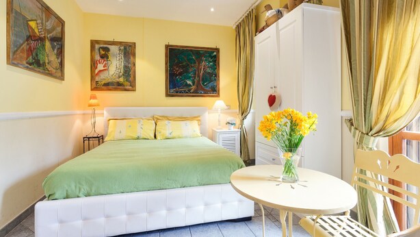 Un dormitorio con cama, mesita de noche con lámpara, una silla, una pequeña mesa con un florero y dos cuadros en la pared.