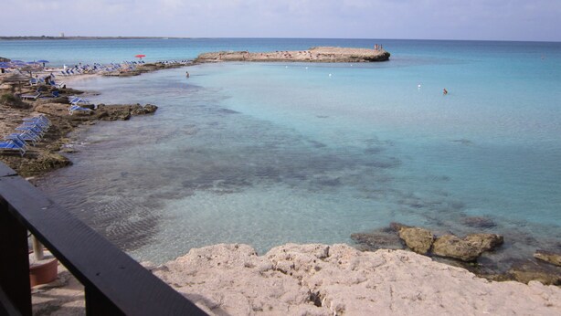 Ein Küstenstrand mit klarem türkisblauem Wasser, felsigem Ufer und Liegestühlen.