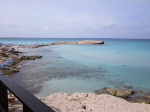 A coastal beach with clear turquoise water, rocky shore, and sun loungers.