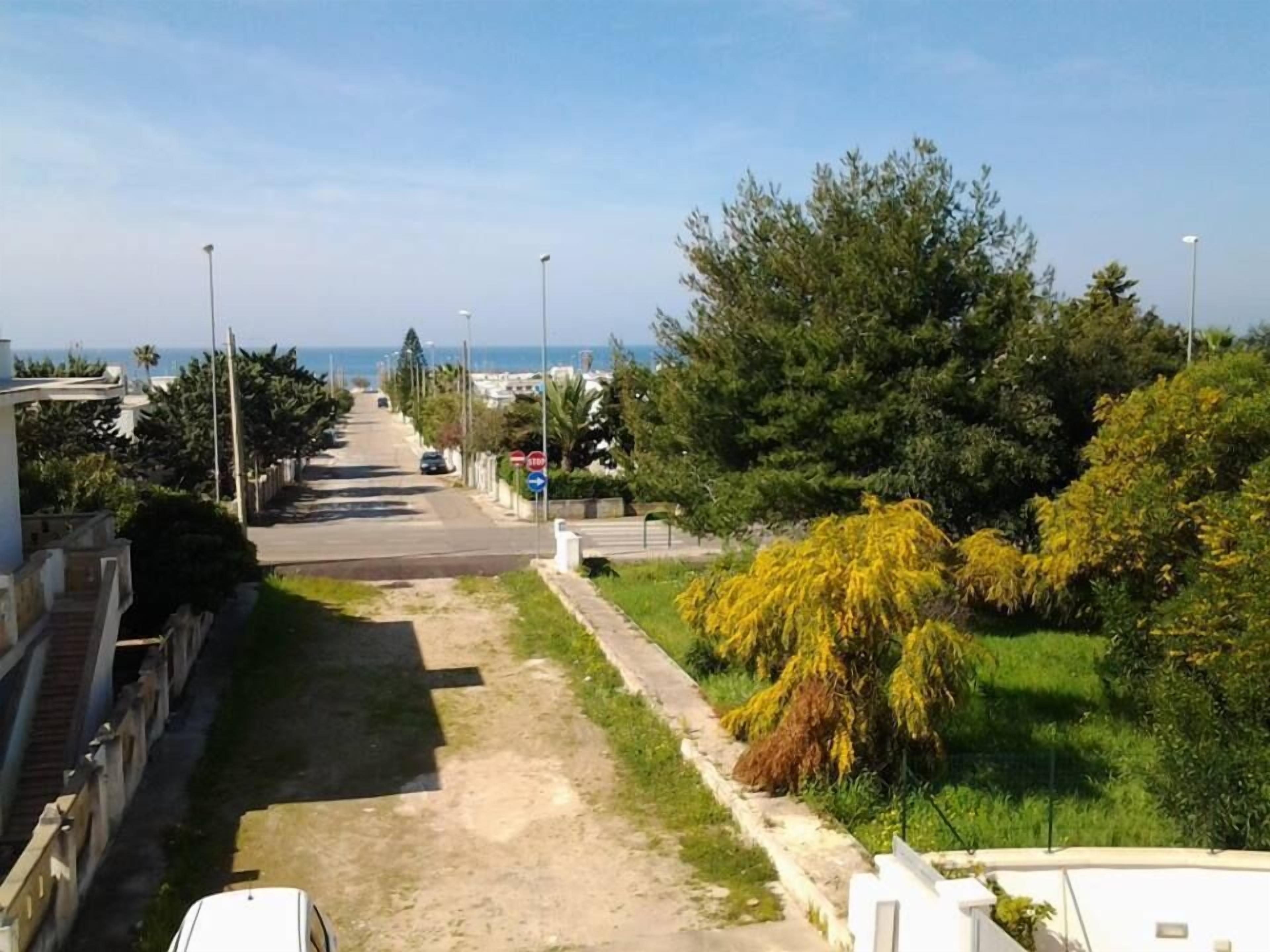 Une rue résidentielle bordée de maisons des deux côtés, avec un trottoir et une vue sur la mer au loin.