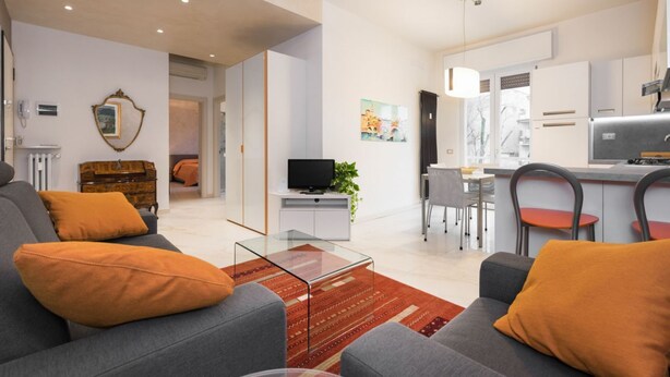 Ein modernes Wohnzimmer mit grauem Sofa, orangefarbenen Kissen, einem Glastisch und einem Fernseher auf einem weißen Schrank. Es gibt eine Essecke mit Tisch und Stühlen sowie eine Küche mit Kühlschrank und Herd.