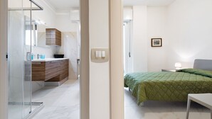 Ein modernes Hotelzimmer mit Duschkabine aus Glas, Holzschränken, einem gesteppten Bett in Grün und einem Nachttisch mit Lampe.