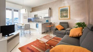 Ein modernes Wohnzimmer mit grauem Sofa, Couchtisch und Fernseher. Es gibt einen Essbereich mit weißem Tisch und Stühlen, sowie eine Küche mit weißen Schränken und einem blauen Wasserkocher.